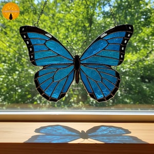 Könnte beinhalten: Ein Buntglas-Schmetterling in Blau- und Schwarztönen, an einer Kette aufgehängt. Die Flügel des Schmetterlings sind aufwendig detailliert und werfen einen Schatten auf die Holzoberfläche. Das Design weist einen Farbverlauf von Blautönen auf.