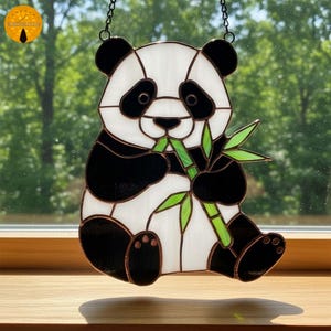 May include: Stained glass panda bear suncatcher with black, white, and green colors. The panda is holding bamboo. The suncatcher is hanging from a chain. The panda bear is a popular animal.