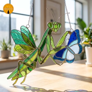 May include: A stained glass suncatcher featuring a green praying mantis with translucent wings, holding a blue and white butterfly. The piece is constructed with dark metal soldering and includes a silver chain for hanging, displaying vibrant colors.
