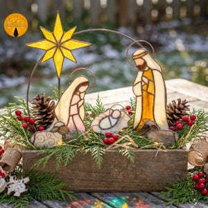 May include: A stained glass nativity scene featuring a yellow star, figures of a parent and child, and a central infant in a manger, all set within a rustic wooden box filled with green pine branches, pinecones, and small red berries.