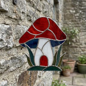 May include: A stained glass mushroom decoration with a red cap, blue accents, and a white stem with a red door. The mushroom is hanging from a metal bracket and chain. The artwork is set against a stone wall.