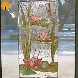 Könnte beinhalten: Buntglasfenster mit einer Seerosenteichszene. Das Kunstwerk zeigt rosa und orangefarbene Seerosen mit grünen Seerosenblättern und hohen grünen Schilf. Das Glas hat eine strukturierte, wellenförmige Wirkung, und das Design ist mit einem silbernen Rahmen eingefasst.