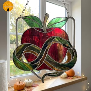 May include: A stained glass suncatcher featuring a deep red apple with green leaves at the top. A long, slender green and amber snake is coiled around the apple in a figure-eight pattern. The piece is framed in metal and hangs by a silver chain.