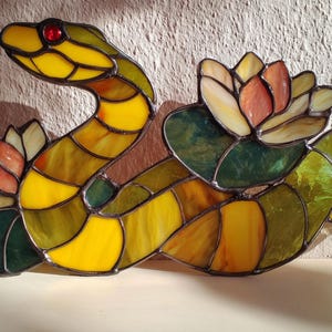 May include: Stained glass sculpture of a yellow and green snake with a red eye, coiled around two water lilies. The lilies have pink, orange, and white petals and green leaves. The artwork is made with various shades of yellow, green, and brown glass.