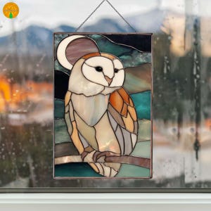 May include: Stained glass panel featuring a barn owl perched on a branch. The owl has a white face, brown wings, and black eyes. The background includes a crescent moon and shades of teal, brown, and green. The panel is framed and hangs from a chain.