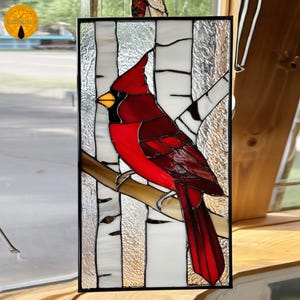 May include: Stained glass panel featuring a vibrant red cardinal perched on a branch. The bird has a yellow beak and black accents. The background depicts birch trees with white and textured glass. The panel is framed in black.