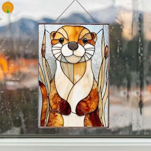 Pode incluir: Painel de vitral com um desenho de lontra. A lontra tem um nariz castanho, olhos pretos e pêlo castanho e branco. O painel inclui juncos e é emoldurado em metal escuro. Uma corrente é fixada para pendurar.