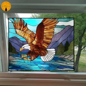 Puede incluir: Panel de vidrieras con un águila calva en vuelo. El águila tiene alas marrones y doradas, cabeza blanca y pico amarillo. El fondo representa montañas, agua y cielo en tonos azules, morados y verdes.
