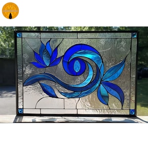 Puede incluir: Panel rectangular de vidrieras con un diseño floral azul. La obra de arte presenta patrones arremolinados y hojas, enmarcados en negro. El fondo transparente realza los vibrantes tonos azules. El panel probablemente se exhibe al aire libre.