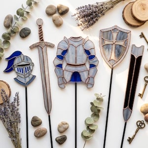 Puede incluir: Un conjunto de cinco estacas de jardín de vitral que incluyen un casco de caballero, una espada, una armadura, un escudo con una cruz y una hoja larga. Cada pieza está hecha con vidrio blanco y azul, soldadura de cobre y varillas de metal.
