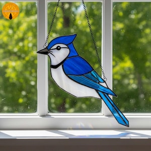 Könnte beinhalten: Glasmalerei-Suncatcher einer Blauhäher, der an einer Kette hängt. Der Vogel hat leuchtende blaue, weiße und schwarze Farben. Der Suncatcher wird vor einem Fenster mit verschwommenem grünem Hintergrund gezeigt.