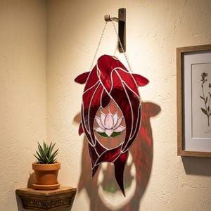 Puede incluir: Un colgante de pared de vidrieras con un pez koi rojo, con una flor de loto rosa en el centro. La obra de arte está suspendida por una cadena de un gancho de bronce. La pieza proyecta una sombra en la pared.