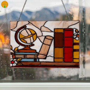 May include: Stained glass panel depicting a library scene with a globe, books, and floral accents. The panel features a rectangular shape with a chain for hanging. The color palette includes shades of brown, red, yellow, and blue, creating a warm and inviting aesthetic.