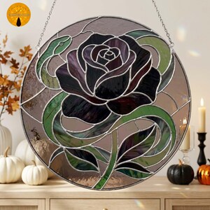 Puede incluir: Un atrapasoles circular de vidrieras con una rosa de color púrpura oscuro en el centro rodeada por una serpiente verde sinuosa. El diseño está compuesto por piezas de vidrio individuales unidas por líneas de soldadura de metal plateado.