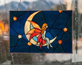 Op de afbeelding: Gebrandschilderd glaspaneel met een fee die op een halve maan zit. De fee heeft oranje haar en kleurrijke vleugels. De achtergrond is diepblauw met steraccenten. Het paneel is ingelijst en hangt aan een ketting.