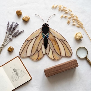 Peut inclure: Un ornement de papillon de nuit en vitrail composé d'ailes translucides crème et beige avec des lignes de soudure brun foncé, un corps central noir uni et de petits accents bleus. Le papillon possède deux fines antennes en fil métallique.
