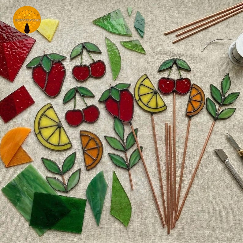 May include: A collection of colorful stained glass fruit and leaf designs, including strawberries, cherries, lemons, and oranges. Some pieces are assembled on copper rods, while others are loose. Various glass pieces and tools are scattered around.