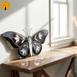May include: A stained glass butterfly wall decor in shades of black, gray, and white. The butterfly features a detailed design with floral accents and is mounted on a white wall. The wings are approximately 24 inches wide.