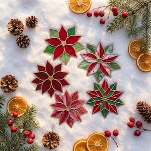 May include: Five stained glass poinsettia ornaments with metal soldering, featuring red and green petals in various patterns. The ornaments are arranged on a white surface with pine cones, dried orange slices, and red berries.