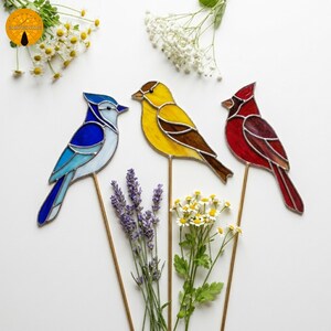 May include: Three stained glass bird garden stakes. A blue jay, a yellow bird, and a red cardinal are displayed. Each bird is mounted on a thin metal rod. The birds are surrounded by white flowers and lavender.