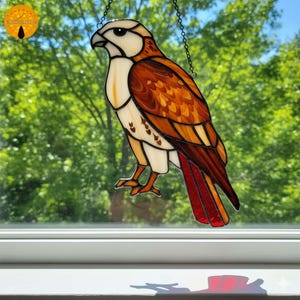 May include: Stained glass hawk with a white head, brown and orange body, and red tail feathers. The bird is hanging from a chain. The background is a blurred view of green trees and a blue sky.
