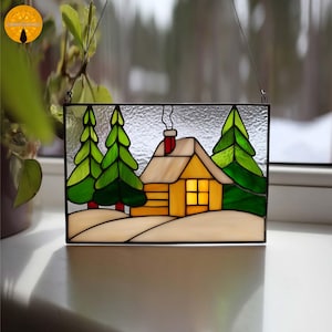May include: Stained glass panel depicting a winter scene with a yellow cabin, green trees, and snow. The cabin has a red chimney and a lit window. The panel is framed in black and hangs by a wire.