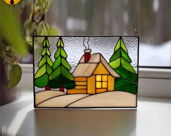May include: Stained glass panel showing a winter scene with a yellow cabin, green trees, and snow. The cabin has a red chimney and a lit window. The panel is framed in black and hangs by a wire.