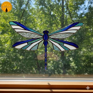 May include: Stained glass dragonfly suncatcher with blue, white, and teal wings. The dragonfly is suspended by a chain and hangs in front of a window with a blurred green background. The dragonfly's body is a deep blue.