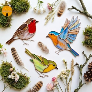 May include: Three stained glass bird ornaments featuring soldered metal edges. One bird has a red breast and brown wings, another is in flight with blue and orange wings, and the third is yellow and green. Each piece includes a small metal hanging loop.