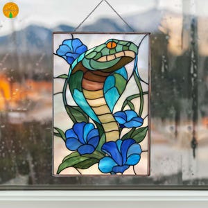 May include: Rectangular stained glass panel depicting a cobra with a green head, brown body, and blue details. The snake is set amongst blue flowers and green foliage. The panel is suspended by a chain.