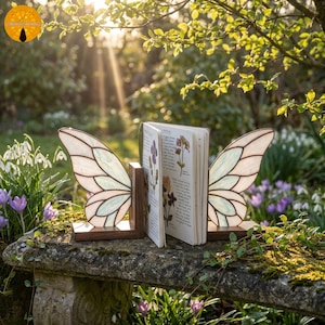 May include: A pair of butterfly wing bookends made of stained glass and wood, holding an open book with pressed flowers. The wings are white, pale green, and cream-colored. The bookends sit on a stone bench in a garden setting.
