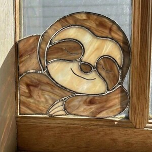 Könnte beinhalten: Buntglasfensterkunst mit einem lächelnden Faultier. Das Faultier ist in Braun- und Cremetönen mit detailliertem Design dargestellt. Das Kunstwerk ist in einem Holzfensterrahmen eingefasst und schafft ein dekoratives Element.