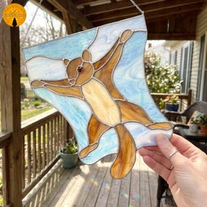 May include: Stained glass panel featuring a brown and cream-colored squirrel with outstretched arms, set against a light blue and white background. The squirrel has a red gem eye and is suspended by a silver chain. A unique, handcrafted art piece.