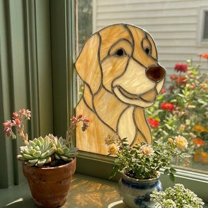 May include: A stained glass window decoration in the shape of a golden retriever dog. The window is set in a green frame, with small potted plants on the windowsill. The plants include succulents and flowering plants.