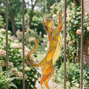 Op de afbeelding: Gebrandschilderde kattenvanger in geel, oranje en goudtinten, hangend aan een ketting. De kat reikt omhoog, met een gekrulde staart. De achtergrond toont een metalen reling en weelderig groen.