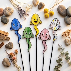 May include: Five stained glass ornaments on thin black metal rods. Each ornament features a unique fish-like shape with a distinct facial expression: a winking blue fish, a smug green fish, a laughing yellow fish, a pink fish with tongue out, and a white fish with colorful swirls.