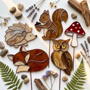 Op de afbeelding: Een verzameling glas-in-loodfiguren waaronder een egel, een eekhoorn met een eikel, een rood-witte paddenstoel, een slapende vos en een uil. Verschillende figuren zitten op dunne metalen stokjes, gerangschikt met verspreide stenen, gedroogde lavendel en varenbladeren.