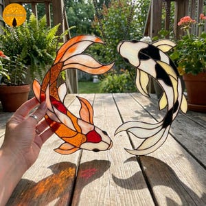 Peut inclure: Deux poissons koï en vitrail suspendus à des chaînes. L'un est orange, rouge et blanc, l'autre noir, blanc et jaune. Les poissons sont présentés sur un fond de verdure et une terrasse en bois.