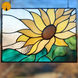 May include: Stained glass panel featuring a vibrant sunflower design. The sunflower has yellow petals, a brown center, and green leaves. The panel is framed in black and hangs from a chain.