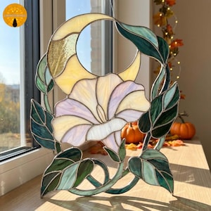 May include: A stained glass suncatcher featuring a central pale lavender morning glory flower surrounded by dark green leaves and a crescent moon shape at the top. The piece is constructed with soldered metal lines and translucent glass panels.