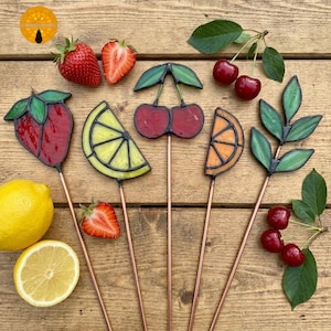 May include: A collection of colorful, stained-glass fruit garden stakes, including a strawberry, lemon slice, cherry, orange slice, and leaves. Fresh strawberries, cherries, and a lemon are also present, arranged on a wooden surface.