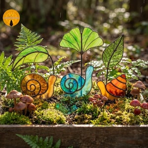 Op de afbeelding: Drie slaksculpturen van glas-in-lood met metalen frames, elk met een unieke kleur huisje: amber, blauwgroen en oranje. Elke slak is bekroond met een groen glazen blad of waaiervormig blad, tentoongesteld tussen kleine bruine paddenstoelen.