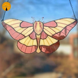May include: A stained glass butterfly suncatcher with a delicate design. The wings feature a blend of pink, yellow, and brown hues, with a detailed pattern. The butterfly is suspended by a chain, perfect for hanging in a window or on a wall.