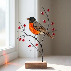 May include: A stained glass robin figurine perched on a dark metal branch with small red glass berries. The bird features a bright orange breast, gray wings, and a black head. The entire structure is mounted on a rectangular wooden base.