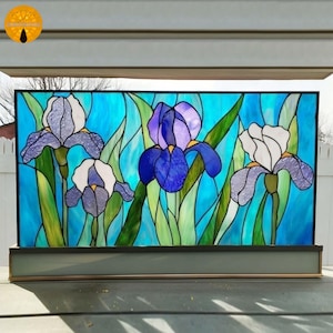 以下が含まれることがあります： 青い背景に、紫と白の色合いの3つのアヤメの花が描かれたステンドグラスパネル。花には緑色の茎と葉があります。パネルは黒いフレームで囲まれ、灰色のベースの上に置かれています。