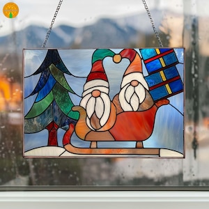 Puede incluir: Panel de vidrieras con dos gnomos en un trineo rojo, con un árbol de Navidad y una caja de regalo. Los gnomos llevan gorros rojos y verdes y tienen largas barbas blancas. El panel está enmarcado y cuelga de una cadena.