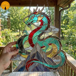 May include: Stained glass dragon suncatcher in teal, red, and gold. The dragon design includes a mountain backdrop and is suspended by a chain. The artwork is displayed outdoors.