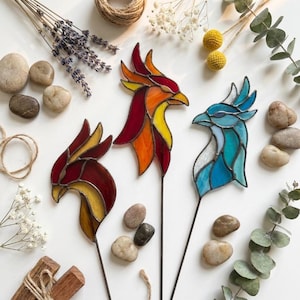 May include: Three stained glass bird head ornaments on thin metal stakes. One bird features deep red and yellow glass, another displays vibrant orange, red, and yellow tones, and the third is crafted from various shades of blue and white glass.
