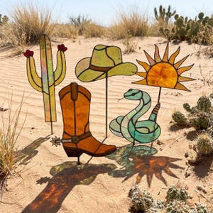 May include: A set of four stained glass garden stakes featuring a yellow cactus with red flowers, a light green cowboy hat, an orange cowboy boot with a silver spur, and a teal snake positioned beneath a bright yellow sun with radiating rays.