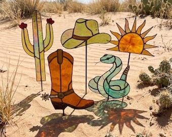 May include: A set of four stained glass garden stakes featuring a yellow cactus with red flowers, a light green cowboy hat, an orange cowboy boot with a silver spur, and a teal snake positioned beneath a bright yellow sun with radiating rays.